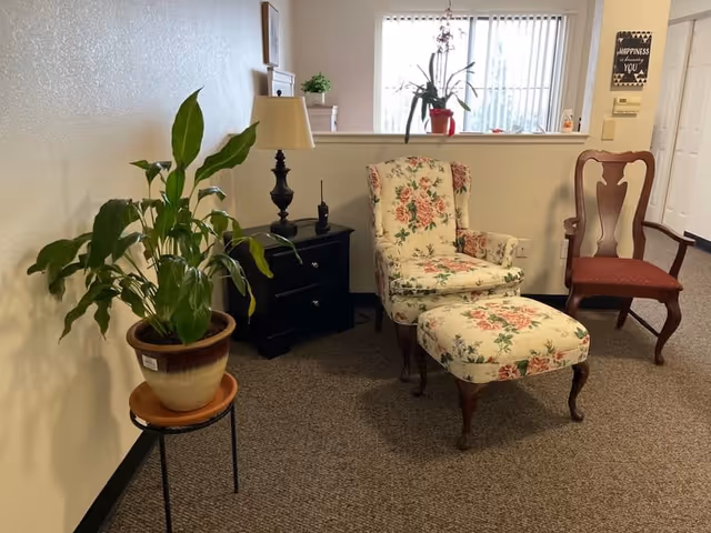 Cozy seating area with a floral armchair and matching ottoman, side table with lamp, potted plants, and wooden chairs in a carpeted interior space.