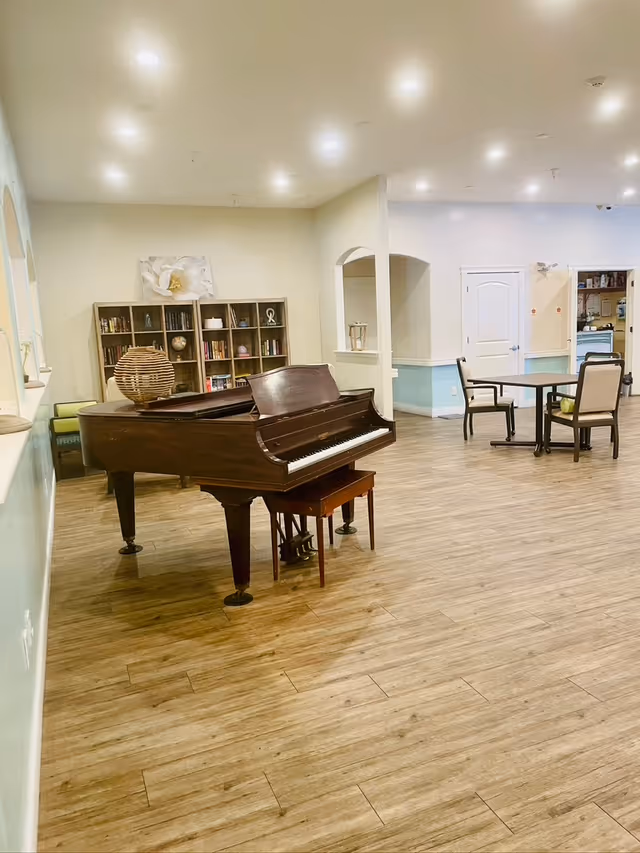 A spacious room with wooden flooring featuring a grand piano with a small bench in the foreground. In the background, there is a bookshelf filled with books and decorative items, a table with four chairs, and a doorway leading to another area. The walls are painted in light colors with a blue accent on the lower half.