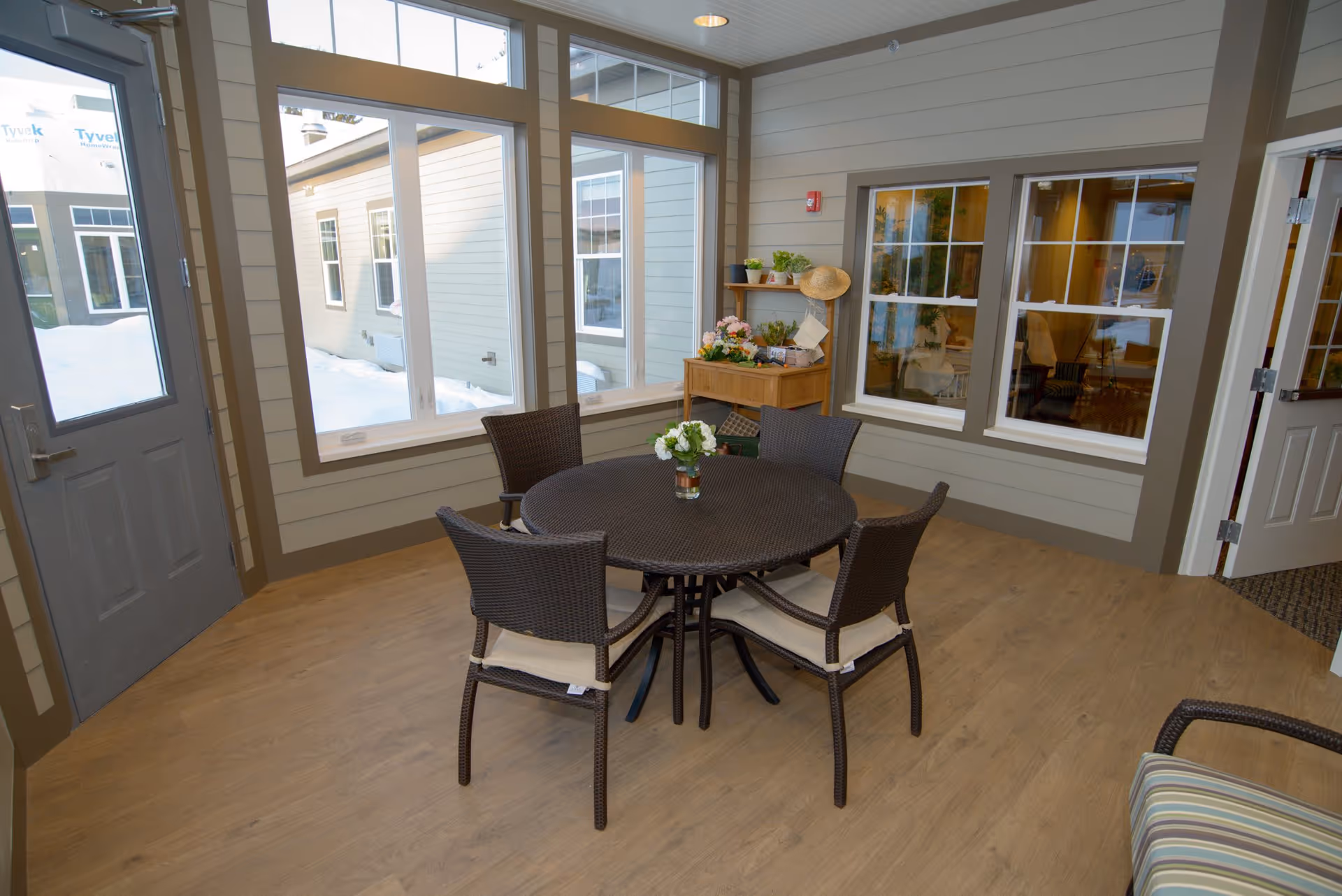 A cozy indoor seating area with a round table and four wicker chairs with cushions. The room has large windows showing a snowy outdoor scene and beige walls with white trim. A small wooden shelf with plants and decorative items is placed against the wall.