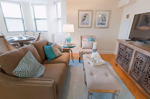 A cozy living room with a brown sofa adorned with patterned and solid teal pillows, a beige cushioned bench coffee table with decorative items, a patterned armchair, a side table with a lamp, two framed bird pictures on the wall, and a wooden TV cabinet with a flat-screen TV. In the background, a dining area with a round table and chairs is visible near large windows with blinds.