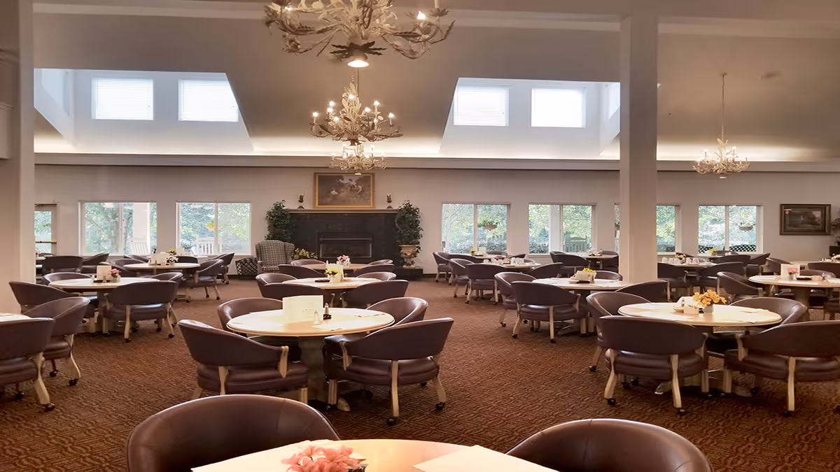 Spacious dining room with multiple round tables and cushioned chairs arranged neatly. The room features large windows letting in natural light, a fireplace with decorative plants on either side, and elegant chandeliers hanging from the ceiling.