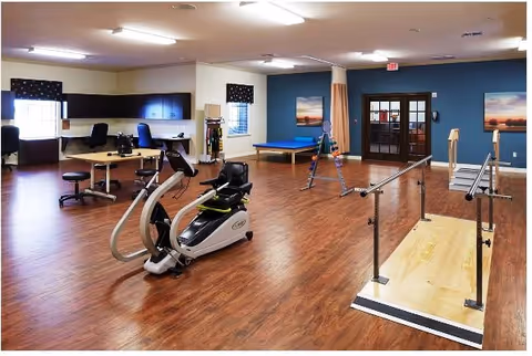 A spacious rehabilitation room with wooden flooring featuring exercise equipment including a stationary bike and parallel bars for walking practice. The room has desks and chairs along the walls, a blue padded therapy table, and large windows with dark valances. The walls are painted in neutral tones with one accent wall in blue, decorated with landscape paintings. There is a door with glass panels and an exit sign above it.