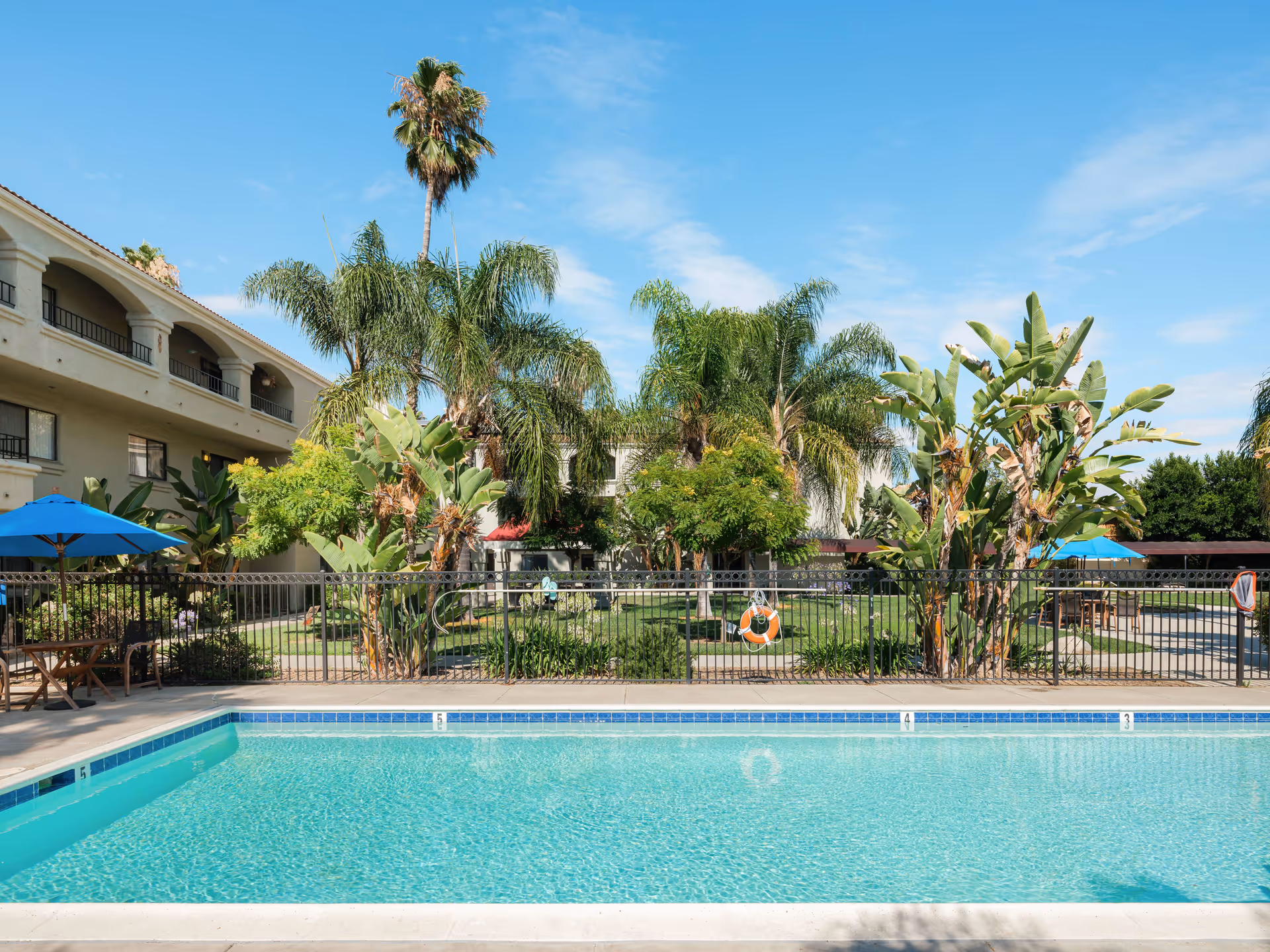 Outdoor swimming pool with clear blue water surrounded by a concrete deck. Behind the pool is a black metal fence, tropical plants including palm trees, and a two-story building with balconies. There are blue umbrellas and wooden tables with chairs on the pool deck. The sky is clear with a few clouds.