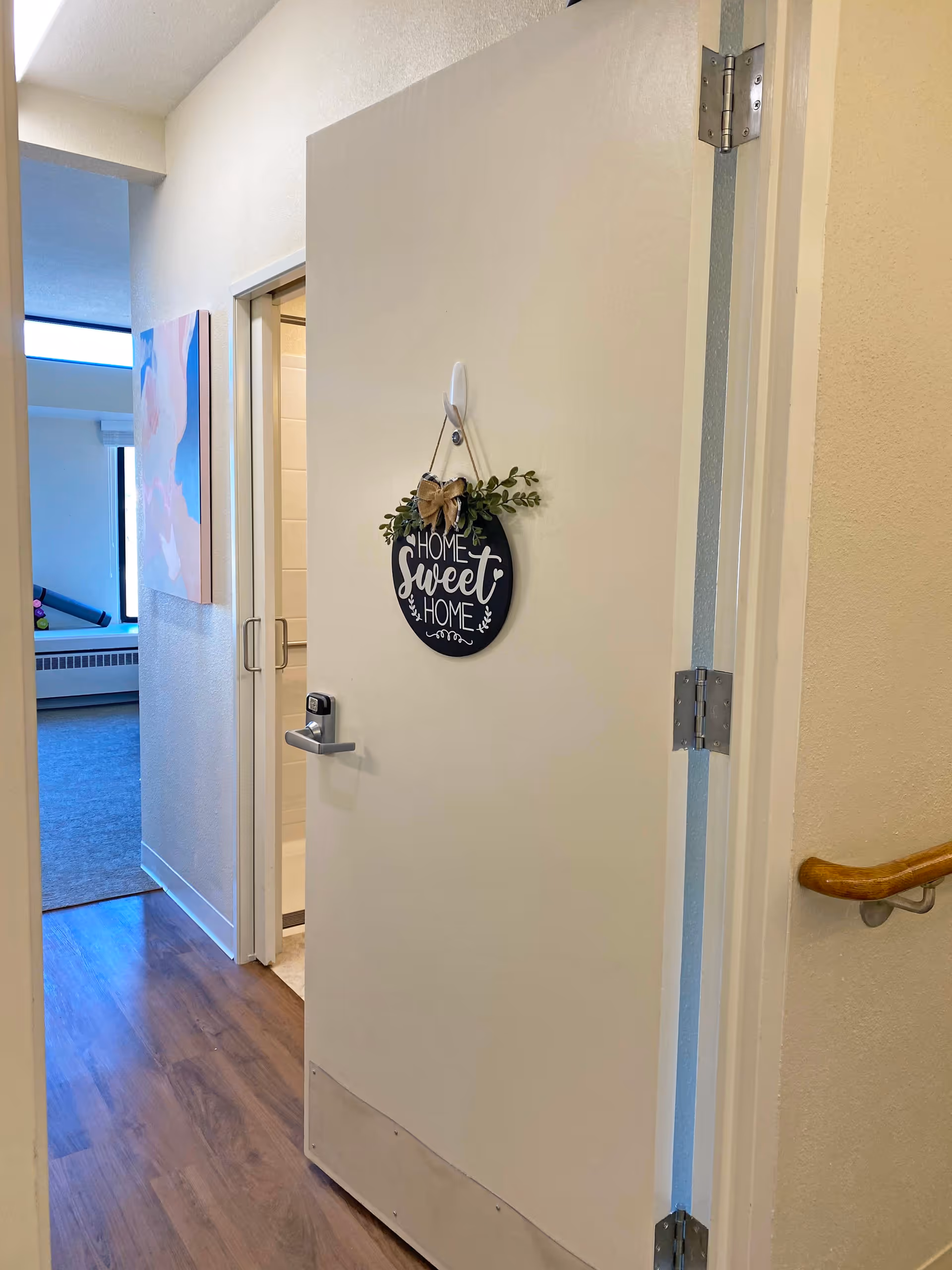 View of an open door with a decorative sign that reads 'Home Sweet Home' hanging on it. The door leads to a bathroom with a glass shower door visible. To the left, there is a hallway with a painting on the wall and a window letting in natural light. The floor is wood, and a handrail is mounted on the wall to the right.