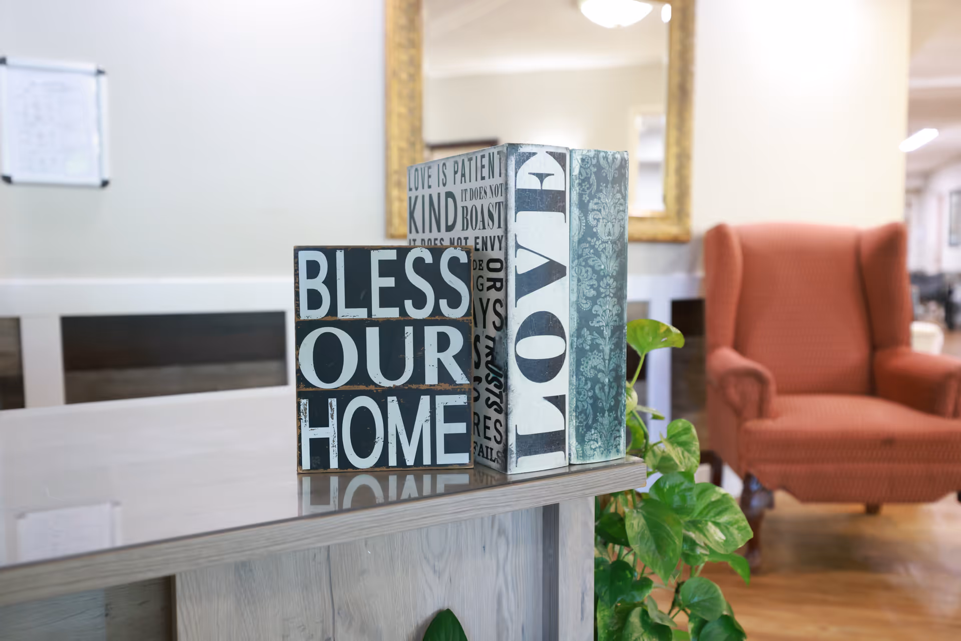 Decorative wooden blocks reading 'BLESS OUR HOME' and 'LOVE' on a reception desk with a red upholstered armchair and plant in the background.