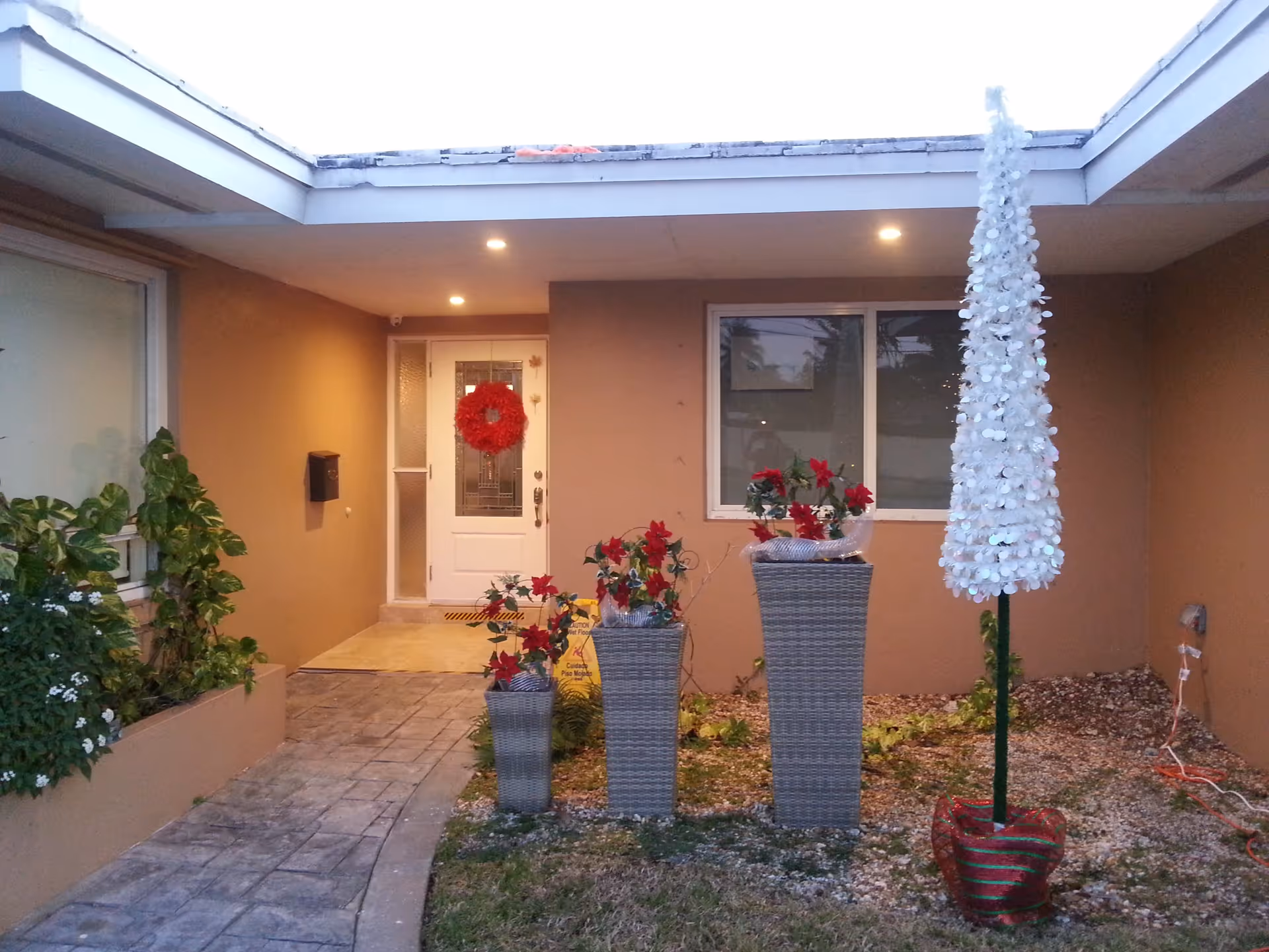 Entrance area of Kendall Gardens Assisted Living Facility decorated for the holidays with a red wreath on the white front door, three tall planters with red poinsettia flowers, and a white artificial Christmas tree in a red and green striped pot. The area is covered with a roof and has warm lighting.
