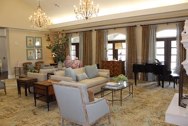 A spacious and elegantly decorated living room with beige sofas, armchairs, a glass coffee table, a grand piano, and large windows with curtains. The room features chandeliers hanging from the ceiling and decorative vases on a side table.