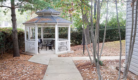 A white wooden gazebo with a shingled roof situated in a garden area surrounded by trees and bushes. There is a concrete pathway leading to the gazebo, which contains a table and chairs inside. The ground is covered with pine needles and leaves.