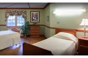 Two single beds in a bright, tidy senior living bedroom with wood floors, a dresser, window with a valance, and a bedside lamp.