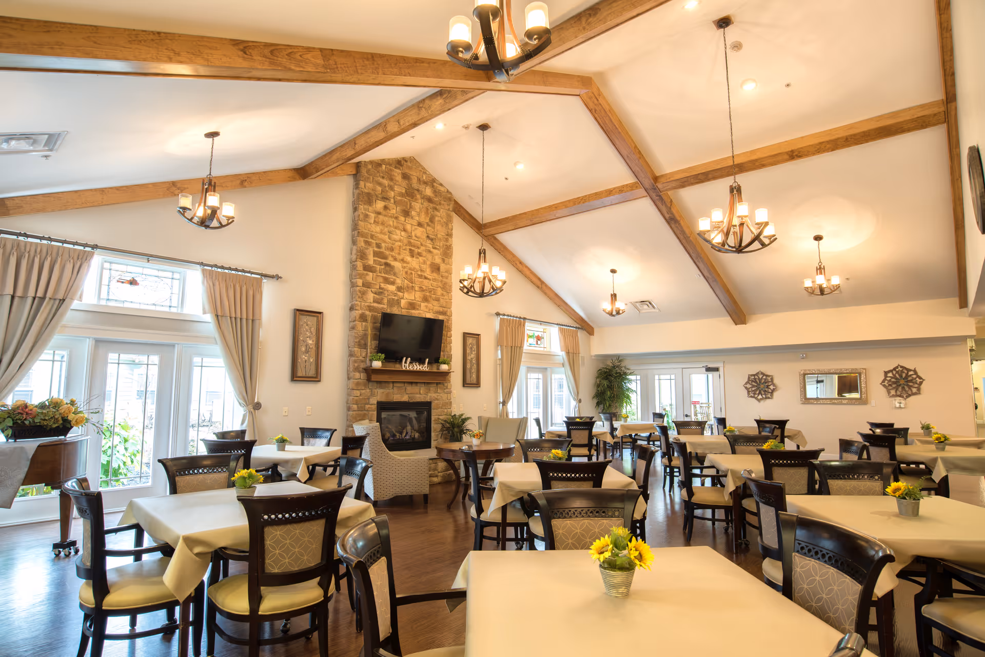 A spacious dining room with multiple tables covered in beige tablecloths, each adorned with small flower arrangements. The room features high vaulted ceilings with wooden beams, several chandeliers, large windows with curtains, a stone fireplace with a mounted TV above it, and comfortable chairs arranged around the tables.