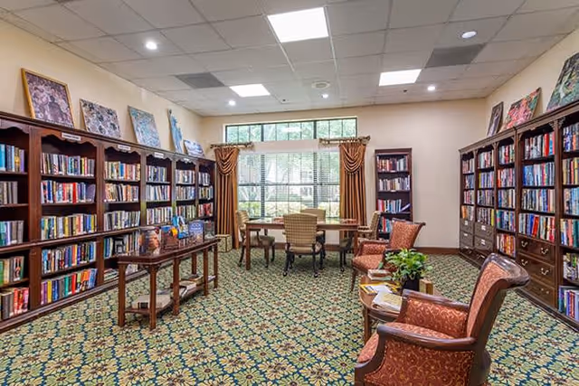 A well-lit library room with multiple wooden bookshelves filled with books along the walls. There are several chairs and tables arranged for reading or studying, including two upholstered armchairs with a small table between them holding a plant and magazines. The room has a patterned carpet and a large window with curtains letting in natural light.