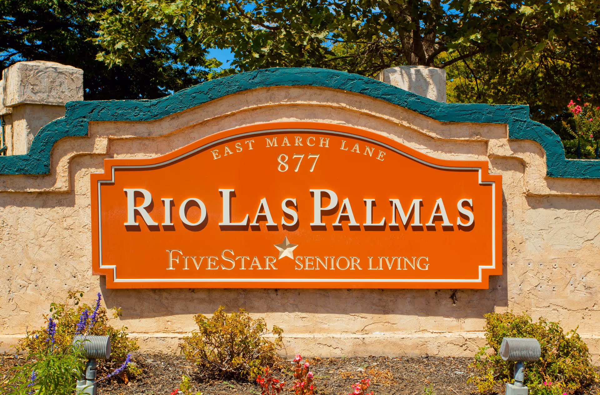 A decorative outdoor sign for Rio Las Palmas Five Star Senior Living located at 877 East March Lane, mounted on a textured beige wall with green trim and surrounded by plants and flowers.
