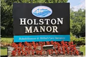 Freestanding sign that reads 'Holston Manor' with smaller text 'Rehabilitation & Skilled Care Services', set among red flowers and greenery.