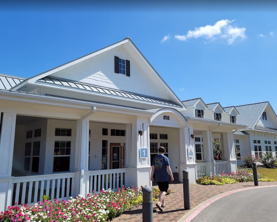 A person walking towards the entrance of a white building with a metal roof and multiple windows. The building has a porch with white railings and columns, flower beds with colorful flowers along the walkway, and the number 818 displayed above the entrance. The sky is clear and blue with a few clouds.
