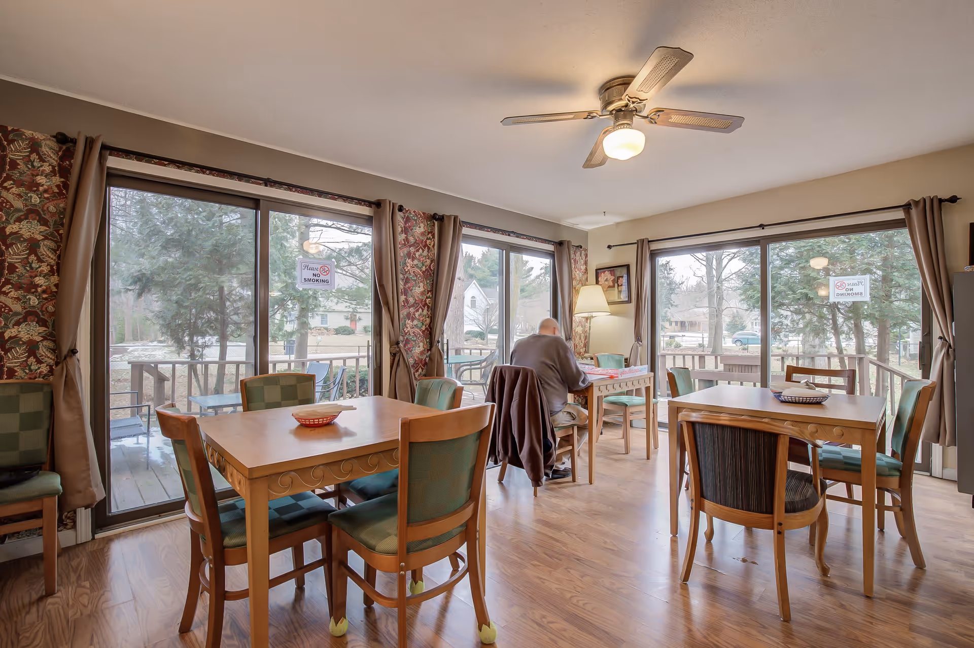 A bright dining room with wooden tables and chairs, large sliding glass doors with curtains, and a ceiling fan. A person is seated at one of the tables near the windows, reading or writing. Outside the windows, there are trees and a deck area. Signs on the glass doors read 'Please No Smoking.'