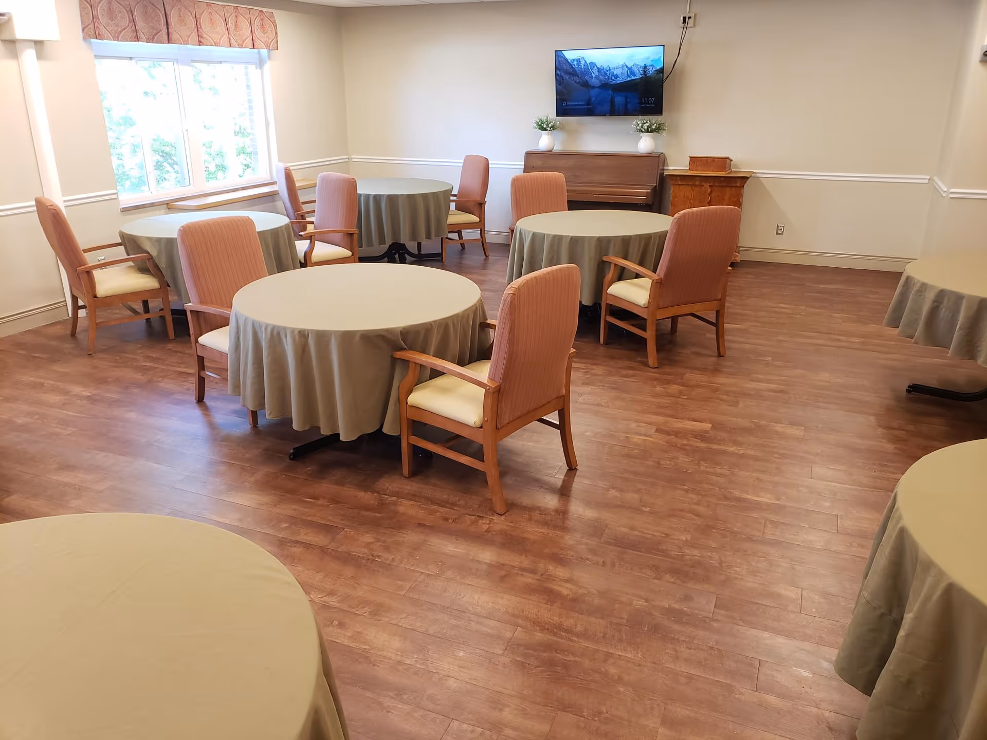 A dining room with several round tables covered with beige tablecloths and surrounded by wooden chairs with cushioned seats and backs. The room has wood flooring, a large window with a patterned valance, and a wall-mounted TV above a piano with decorative plants on top.