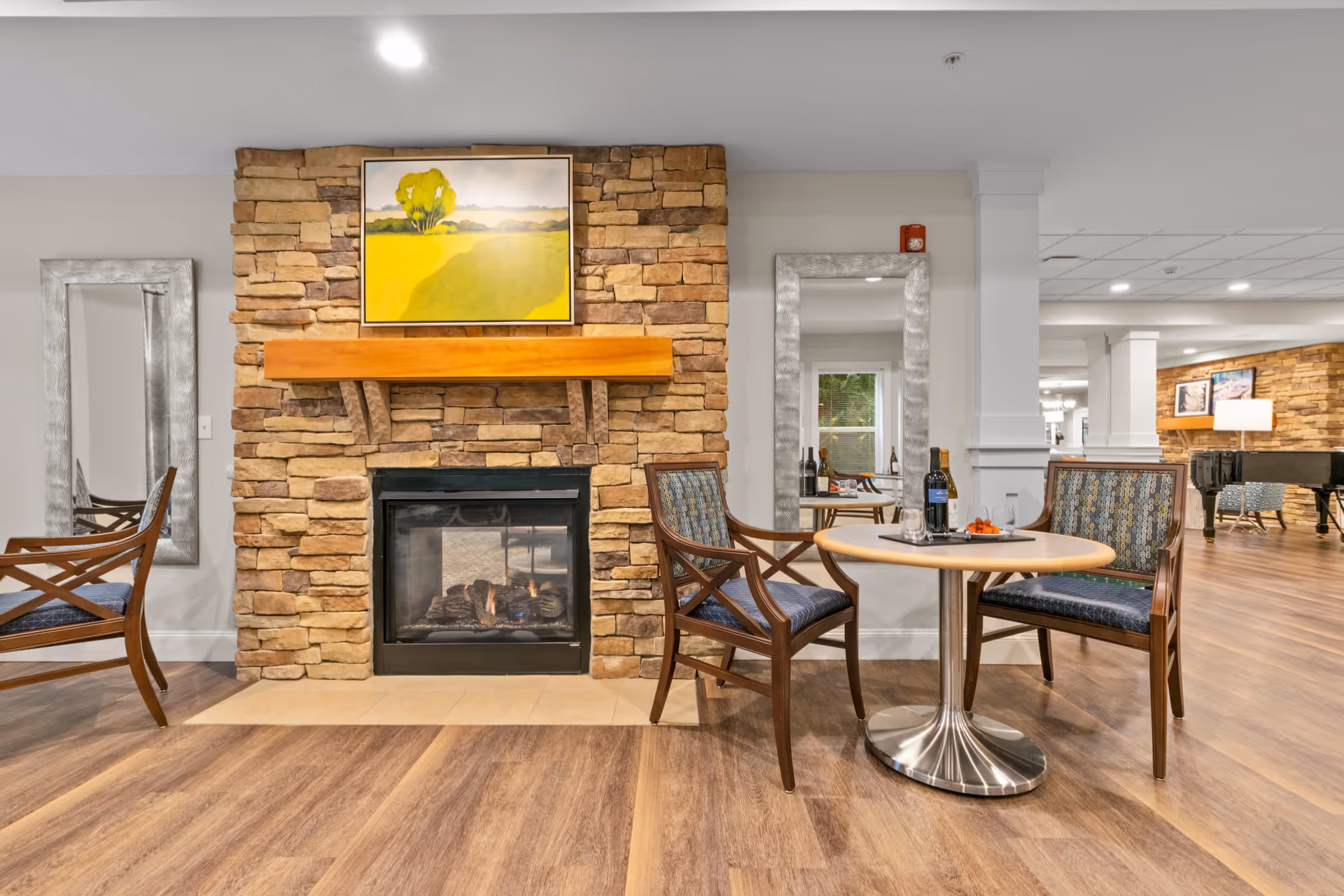 Cozy common area with a stone fireplace, wooden mantel, two chairs and a small round table on wood flooring.