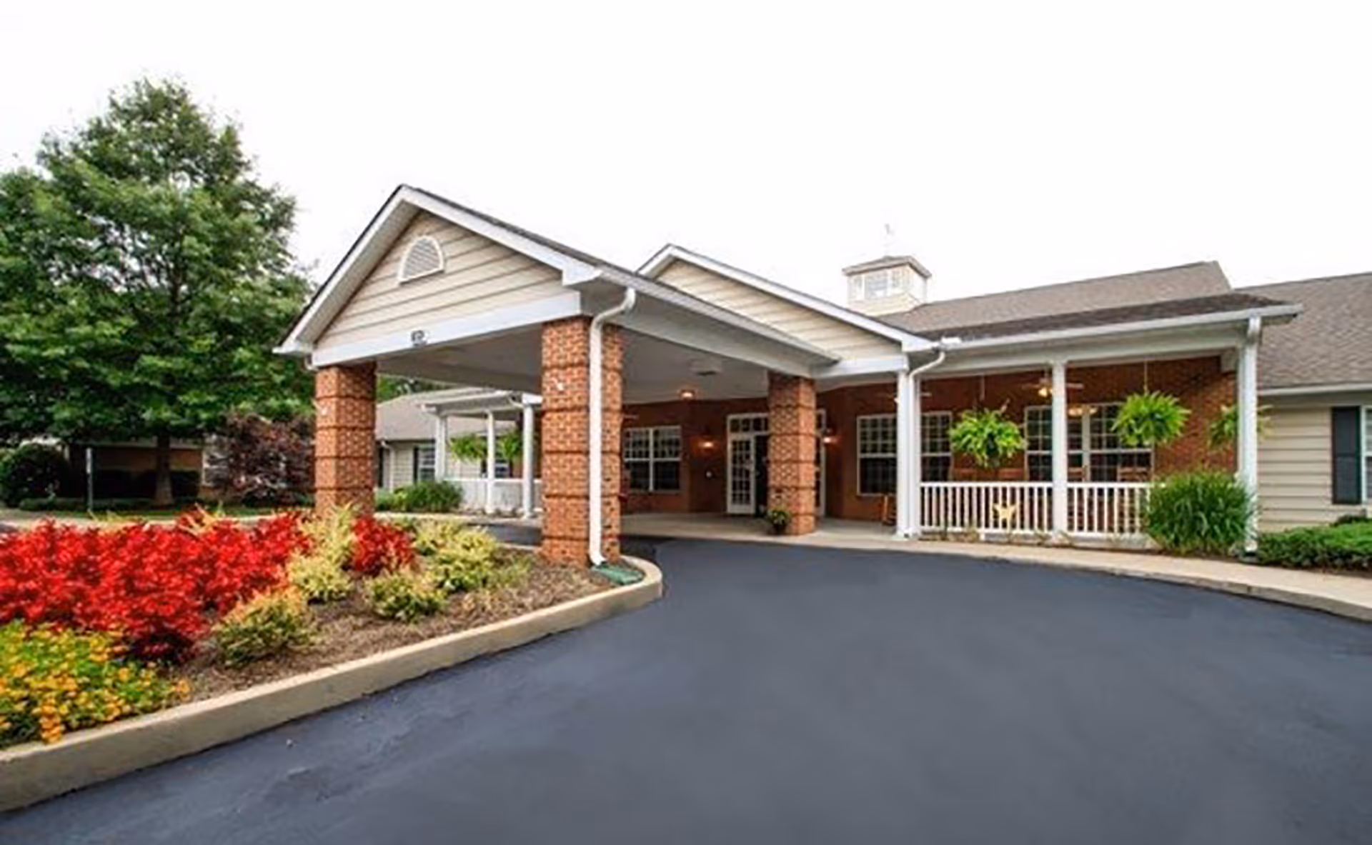 Front exterior view of Spring Arbor Assisted Living facility showing a covered entrance with brick pillars, a driveway, landscaped flower beds with red and yellow flowers, and green trees in the background.