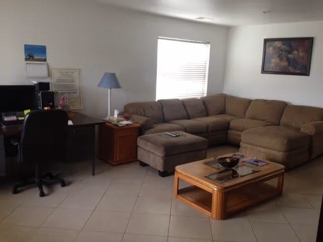 A living room with a large beige sectional sofa and matching ottoman, a wooden coffee table with a glass top, a desk with a computer and office chair, a floor lamp, and a window with blinds letting in natural light. A framed picture hangs on the wall above the sofa.