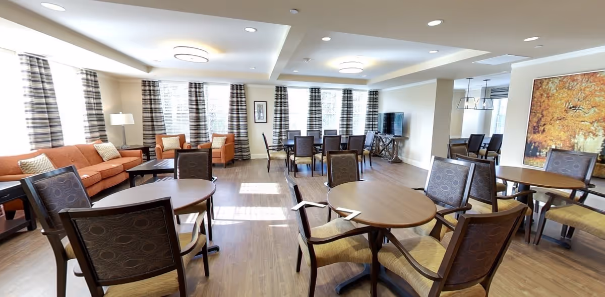 A spacious and well-lit common area with multiple round wooden tables and chairs arranged for seating. There is a cozy sitting area with an orange sofa and matching armchairs near large windows with striped curtains. The room features wood flooring, modern ceiling lights, a flat-screen TV on a stand, and a large framed autumn-themed artwork on the wall.