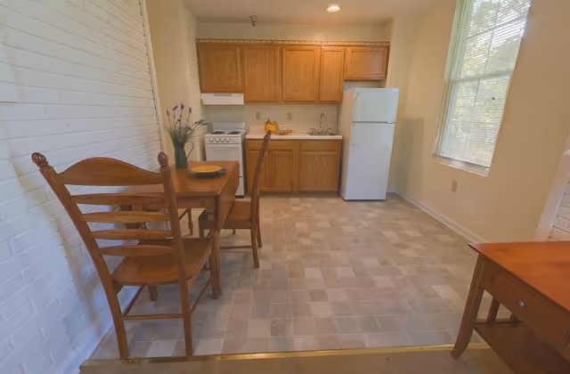 Small kitchenette with oak cabinets, a white stove and refrigerator, and a wooden dining table with chairs beside a window.