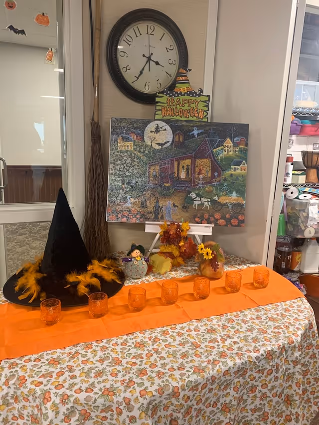 A Halloween-themed display on a table covered with a floral tablecloth. The display includes an orange runner with six orange glass candle holders, a black witch hat decorated with orange feathers, a small scarecrow doll, autumn leaves, and a pumpkin vase with yellow flowers. Behind the table is a completed Halloween puzzle on an easel, a broomstick leaning against the wall, and a round wall clock showing the time as 4:35. The background includes shelves with various craft supplies.