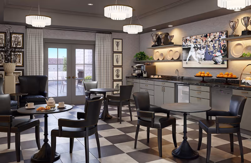 A cozy common area with black leather chairs and round tables on a checkered floor. The room features a kitchenette with a coffee machine, sink, and countertop with bowls of oranges. A flat-screen TV on the wall displays a baseball game. French doors lead to an outdoor patio with seating. The room is softly lit by modern pendant lights and decorated with framed pictures and plants.