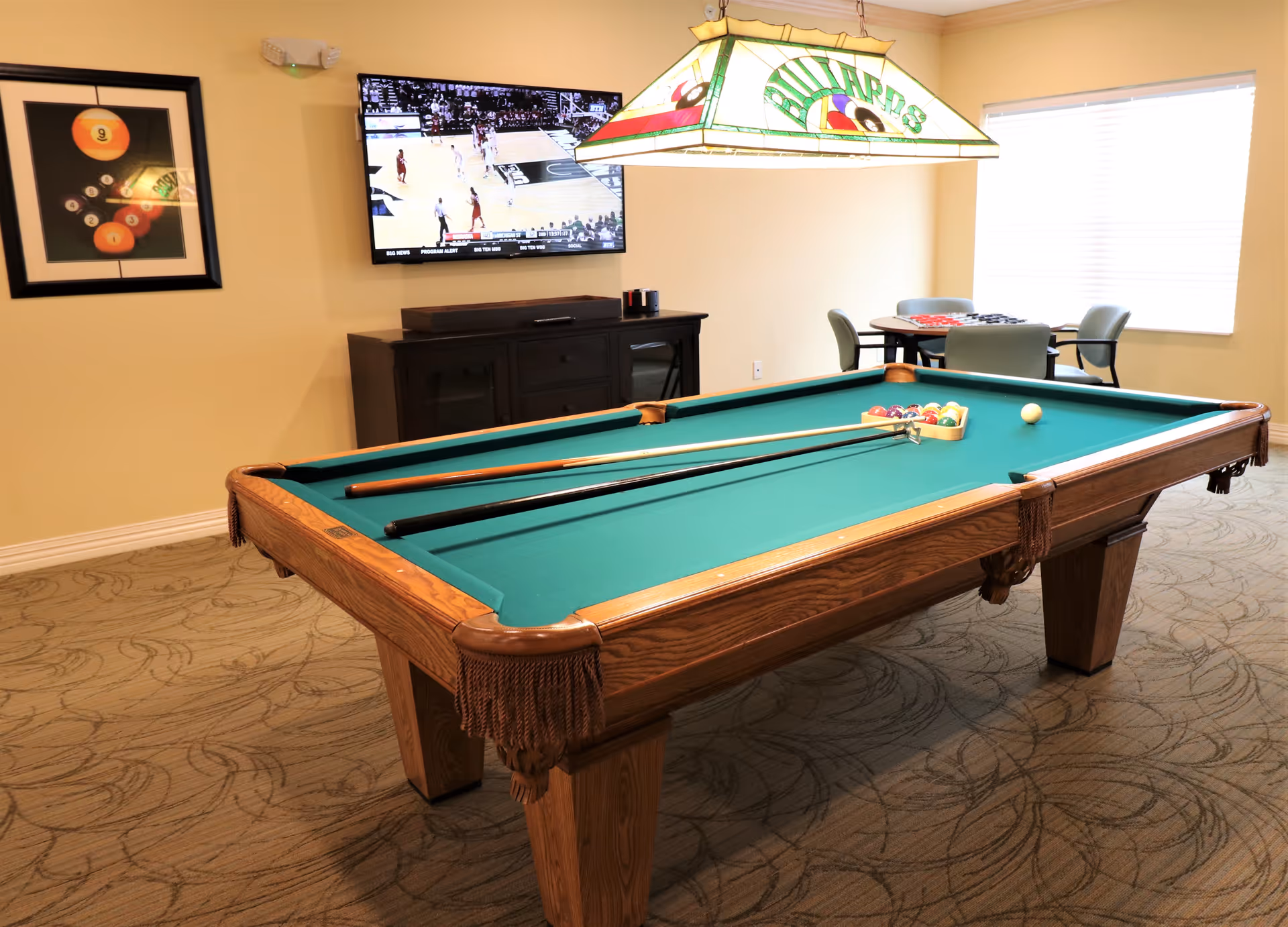 A billiards room featuring a green pool table with cues, a stained-glass hanging light, a wall-mounted TV, and a small table by the window.