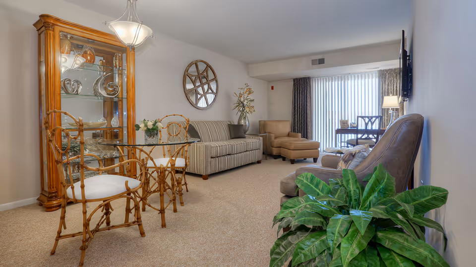 A cozy living room in a senior community featuring a glass-top dining table with two wooden chairs, a striped sofa, a beige armchair with an ottoman, a large green plant, a wooden display cabinet with glass shelves, a round decorative mirror on the wall, and a window with vertical blinds letting in natural light.