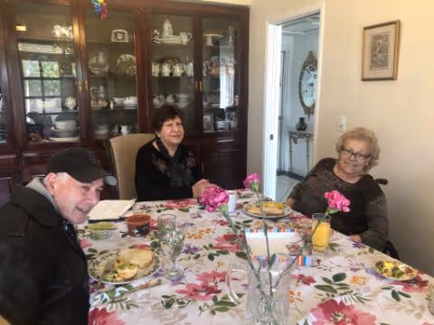 Three seniors seated around a dining table with a floral tablecloth and plates, in a room with a china cabinet.
