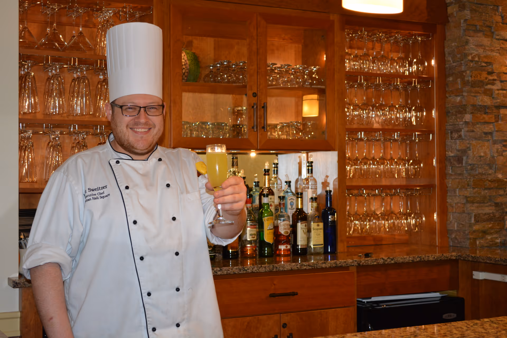 A chef in a white uniform and tall hat holds up a cocktail behind a bar stocked with glassware and bottles.