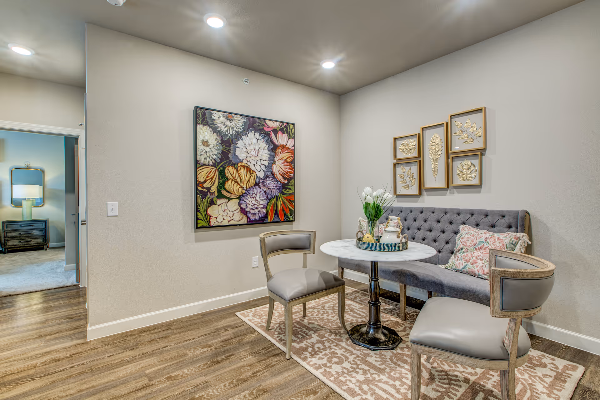 A cozy seating area with a small round marble table, two gray cushioned chairs, and a tufted gray bench with floral pillows. The area is decorated with a floral painting on one wall and four framed botanical prints on another. The floor has a patterned rug and wood-style flooring, with a glimpse of another room in the background.