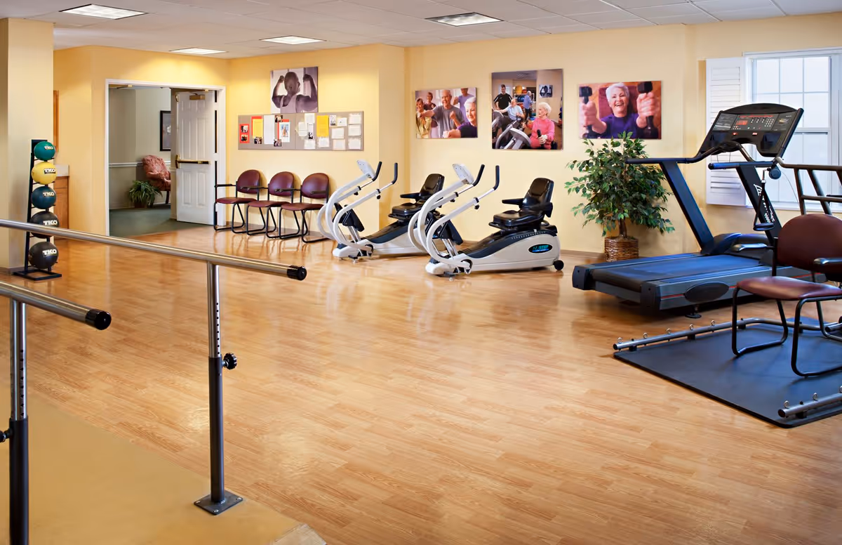 A senior living facility exercise room with wooden flooring, parallel bars for walking support, two recumbent exercise bikes, a treadmill, and a rack of colorful exercise balls. There are four chairs lined up against a yellow wall with bulletin boards and photos of elderly people exercising. A potted plant is placed near the treadmill, and a doorway leads to another room.