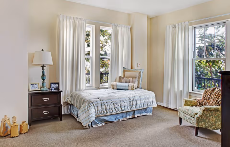 Sunny furnished bedroom with a single bed, nightstand with lamp and framed photos, and an upholstered armchair by large windows with white curtains.