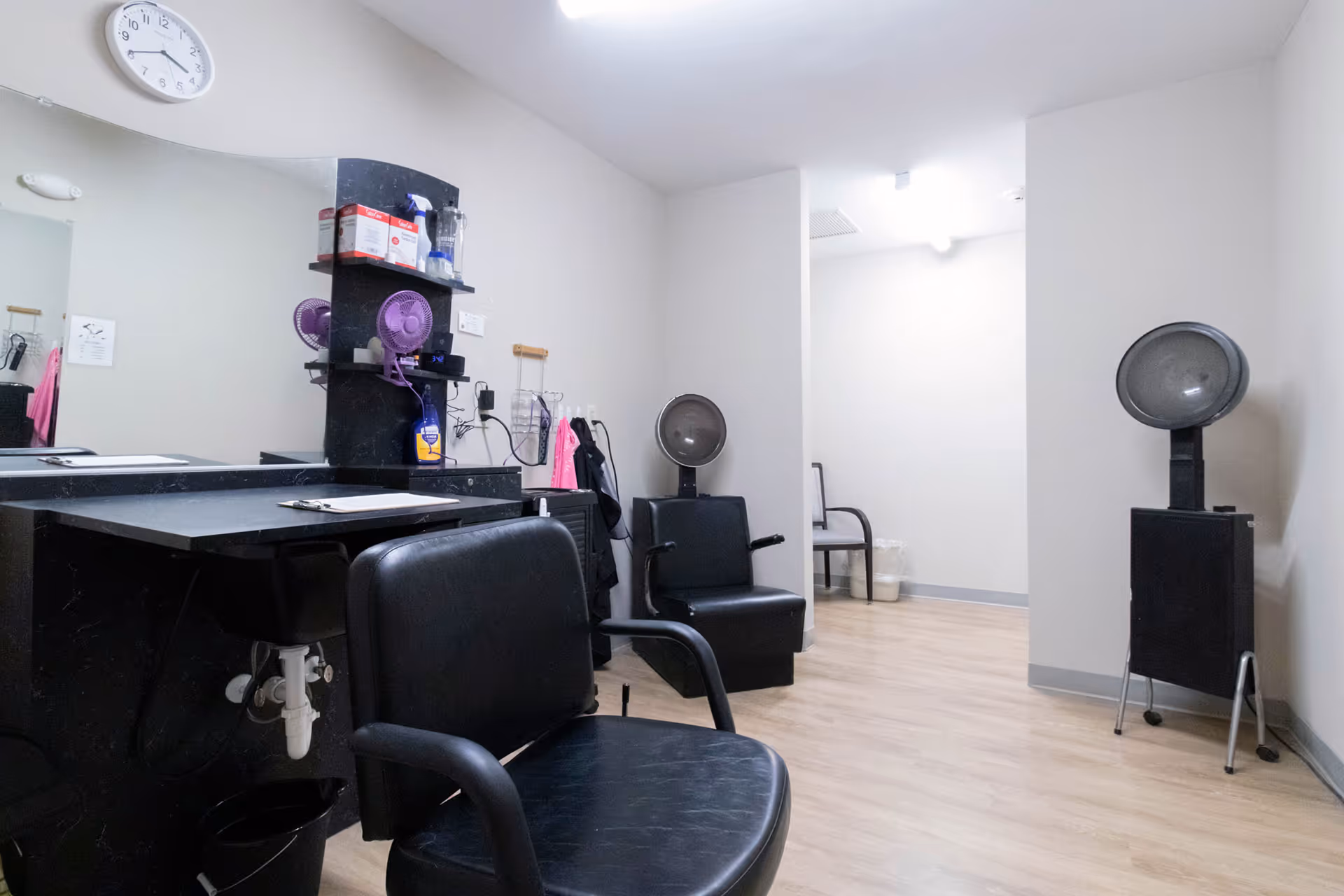 Interior hair salon with black styling chairs, a large mirror, shelves of hair products, and drying stations.