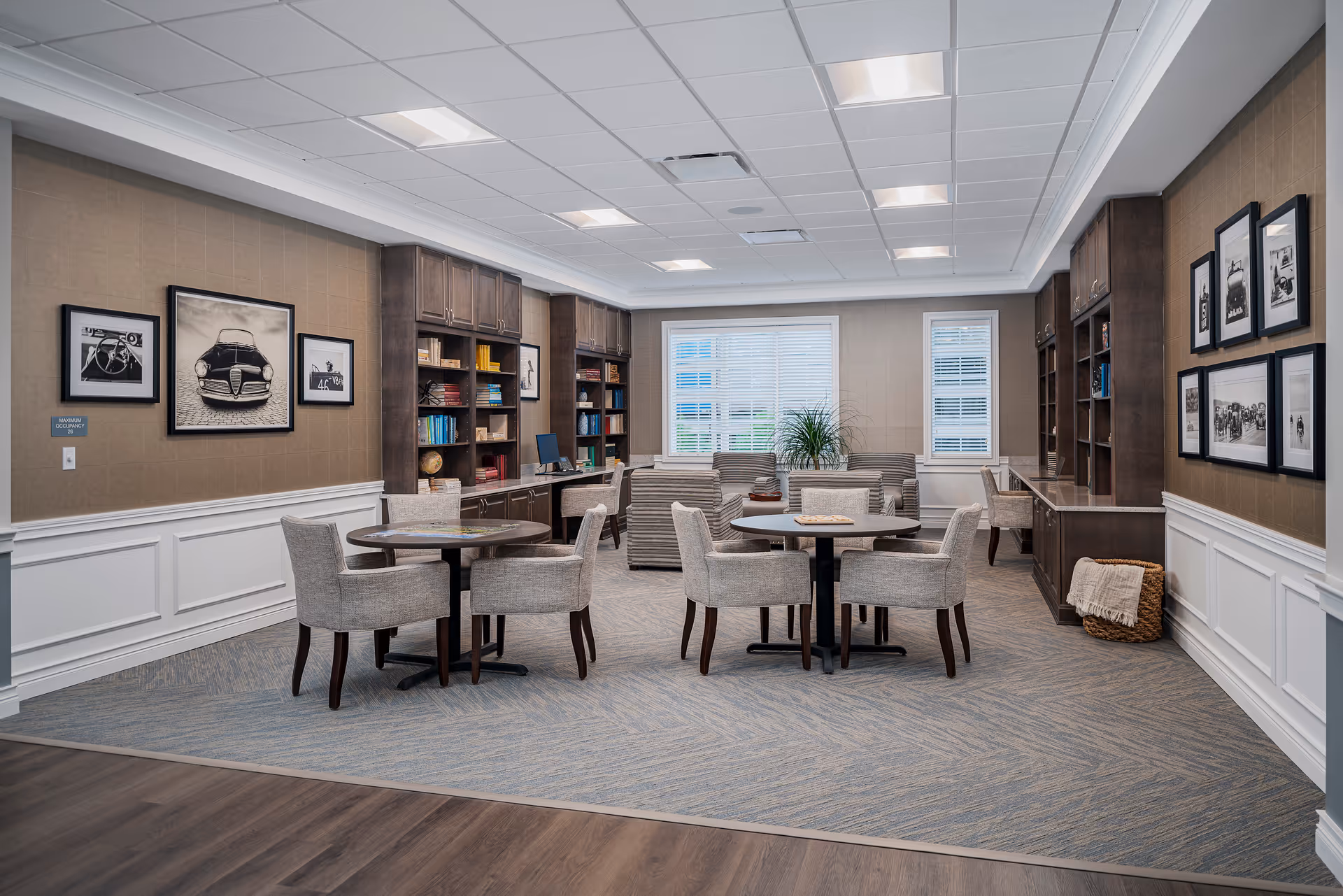 A well-lit interior room with two round tables each surrounded by four upholstered chairs. The room features built-in dark wood bookshelves filled with books and decorative items, a computer on a desk, framed black and white photographs on the walls, and a large window with white blinds letting in natural light. The floor has a combination of wood and carpet, and there is a basket with a blanket in the corner.