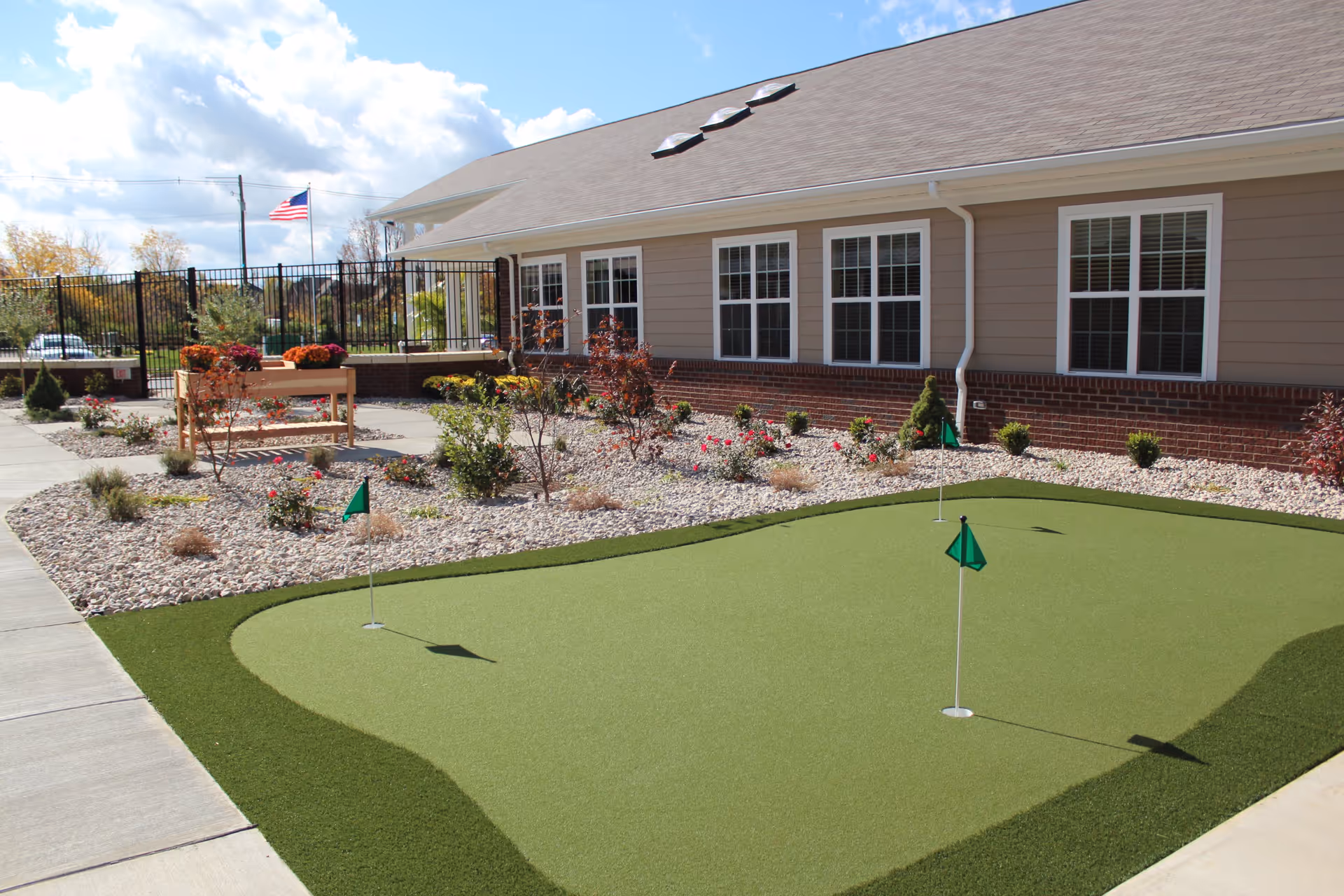 Outdoor putting green with three small green flags in front of a single-story building with multiple windows. There is a landscaped area with rocks, plants, and flowers adjacent to the putting green. An American flag is visible in the background under a partly cloudy sky.
