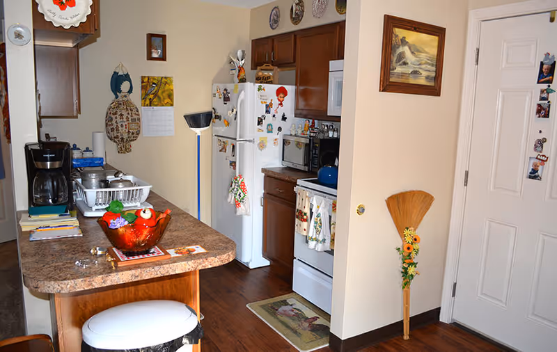Interior view of a kitchen area with wooden cabinets, a white refrigerator covered with magnets, a white stove with oven mitts hanging on the handle, a microwave, and a coffee maker on the counter. There is a countertop with a red decorative bowl and some papers, a broom with flowers leaning against the wall, and a door with photos attached to it. The floor is wooden and the walls are beige with some decorative plates and a painting.