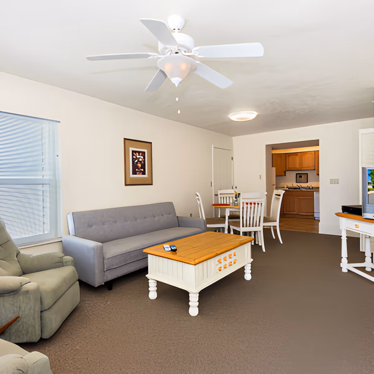 A living room area in a retirement community featuring a gray sofa, a green recliner chair, a wooden coffee table with white legs, a ceiling fan with lights, a small dining table with four chairs, and a view into a kitchen with wooden cabinets. The room has beige walls and carpeted flooring.
