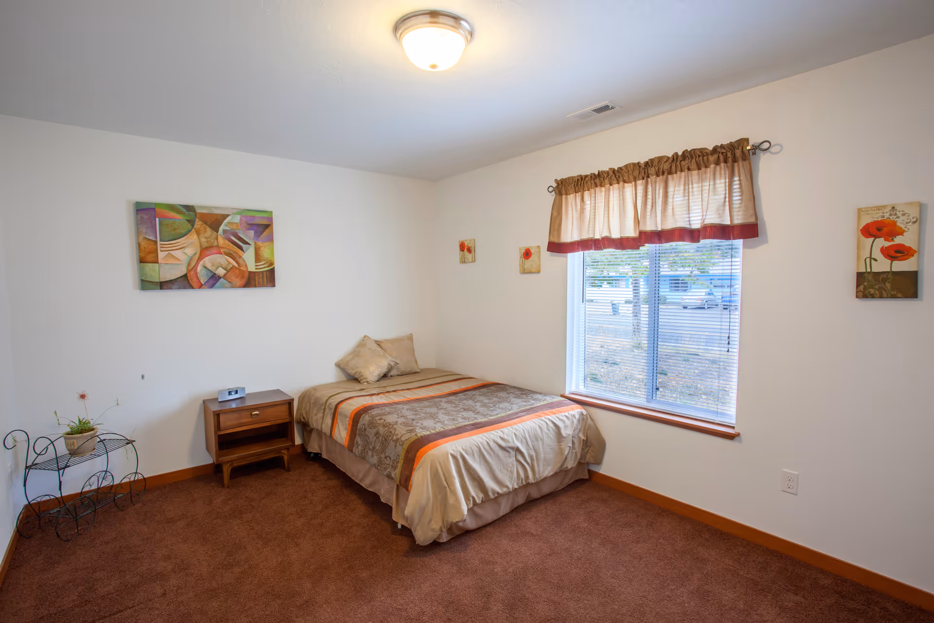Simple furnished bedroom with a single bed, nightstand, window with a valance, wall art, and brown carpet.