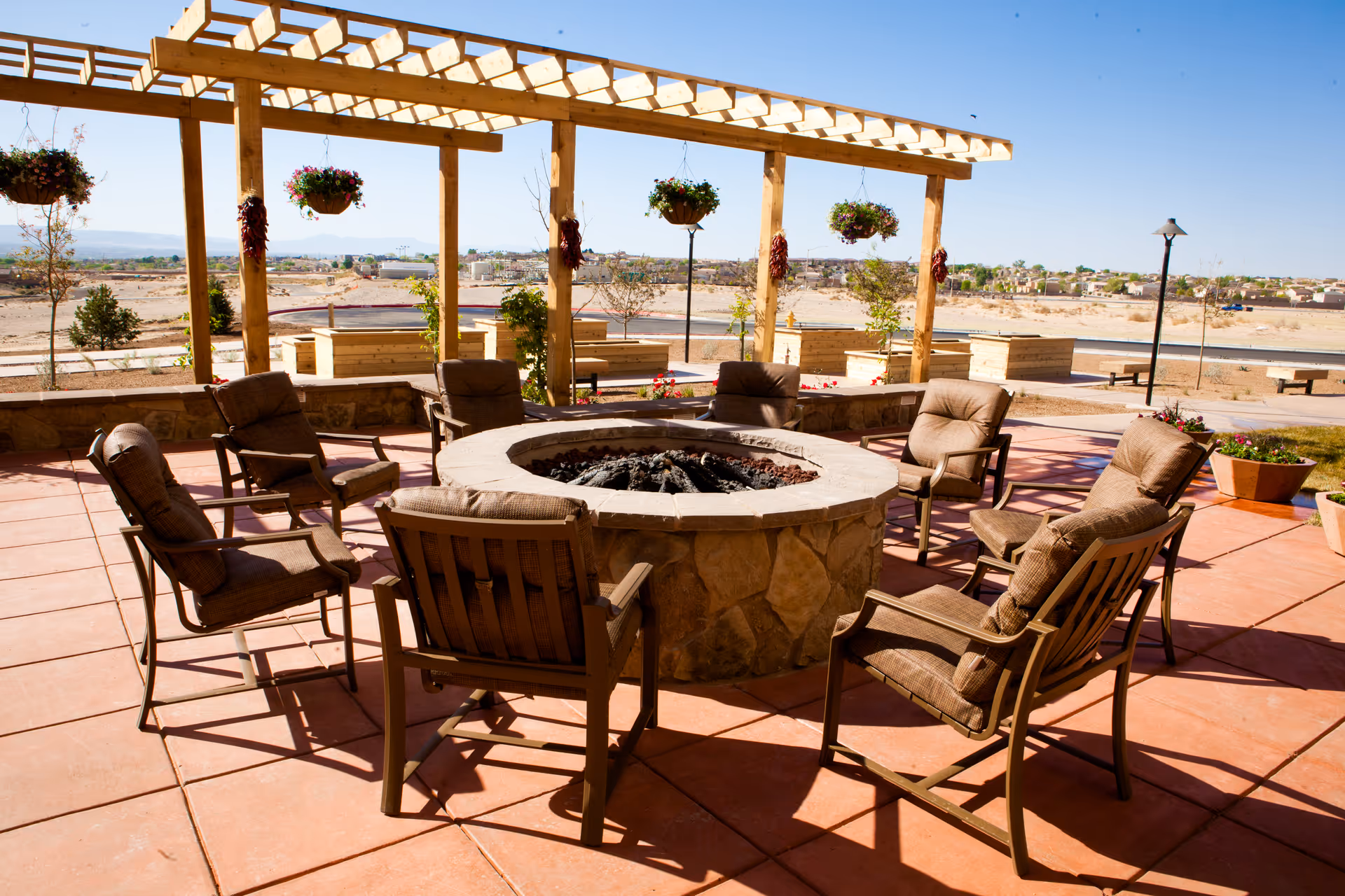Outdoor patio with a circular stone fire pit surrounded by cushioned chairs under a wooden pergola overlooking a desert landscape.