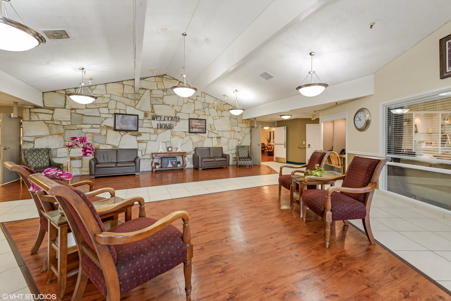A spacious and well-lit common area with wooden flooring and a tiled walkway. The room features several wooden chairs with maroon cushions arranged around small tables, a stone accent wall with framed pictures and a 'WELCOME HOME' sign, two brown leather sofas, and patterned armchairs. Ceiling lights hang from the white ceiling, and a clock is mounted on the wall near a large window looking into another room.