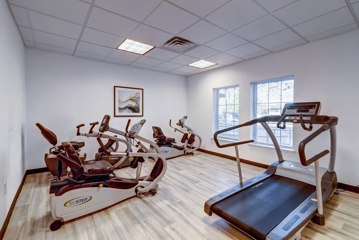 A small fitness room with three recumbent exercise bikes and one treadmill. The room has light wood flooring, white walls, a framed abstract painting, and two windows with blinds letting in natural light.