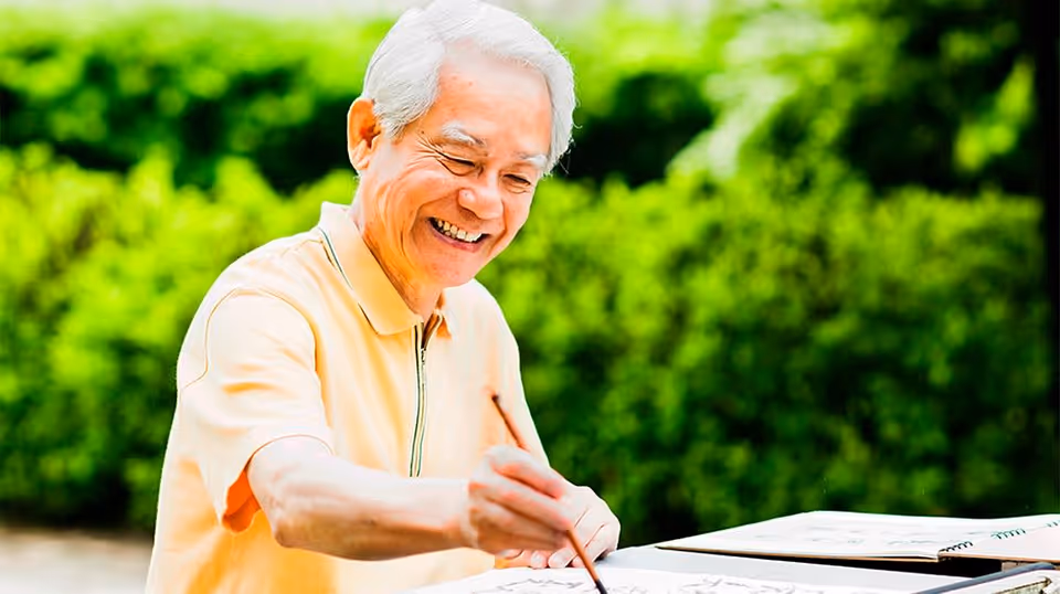 An elderly man with white hair wearing a light yellow polo shirt is smiling and painting outdoors with green bushes in the background.
