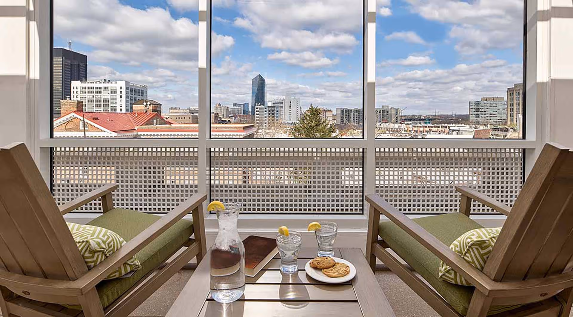 Two wooden lounge chairs with green cushions and patterned pillows face a large window overlooking a cityscape under a partly cloudy sky. A small table between the chairs holds a glass pitcher of water with lemon slices, two glasses of water with lemon, a book, and a plate with cookies.