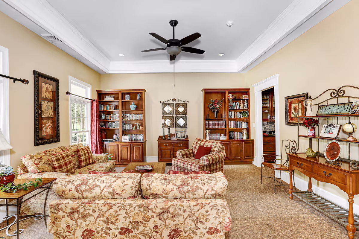 A cozy living room with floral and plaid upholstered sofas and armchair, wooden bookshelves filled with books and decorative items, a ceiling fan, framed artwork on the walls, and a wooden console table with decorative pieces including a 'Welcome Cottage' sign.