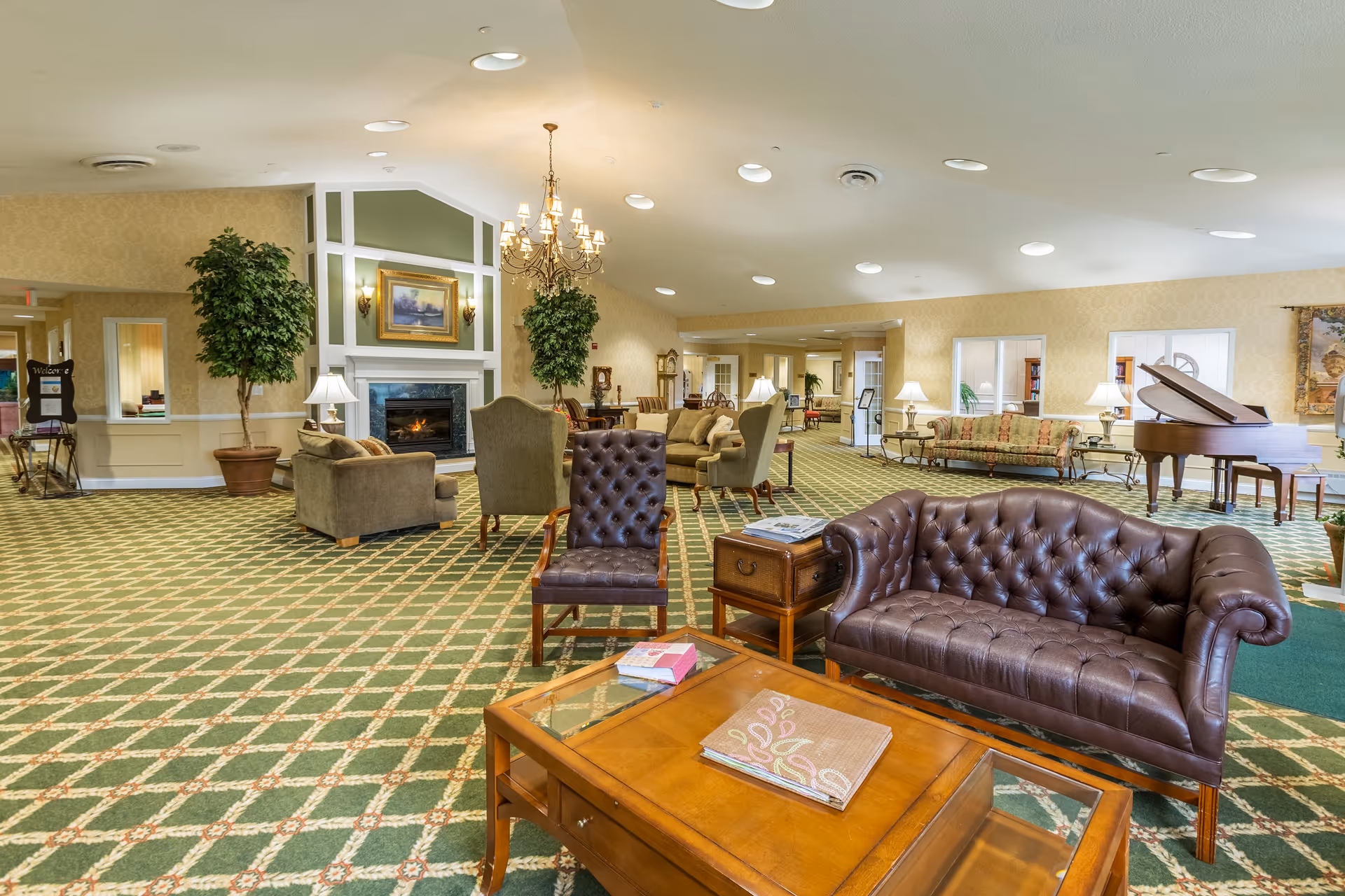 A spacious and well-lit living room area in a senior living facility featuring a green patterned carpet, a brown leather tufted sofa, a matching armchair, a wooden coffee table with books, several other seating arrangements including sofas and armchairs, a grand piano, large potted plants, a chandelier, and a fireplace with a framed painting above it.