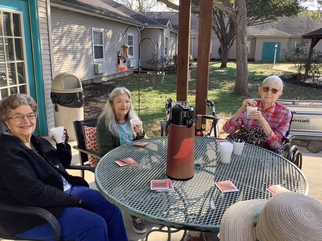 Three elderly women sitting around a round metal outdoor table with playing cards and white cups, enjoying drinks and smiling. They are outside near a building with beige siding, and there is a tree and some greenery in the background. One woman is in a wheelchair, and a large thermos is on the table.