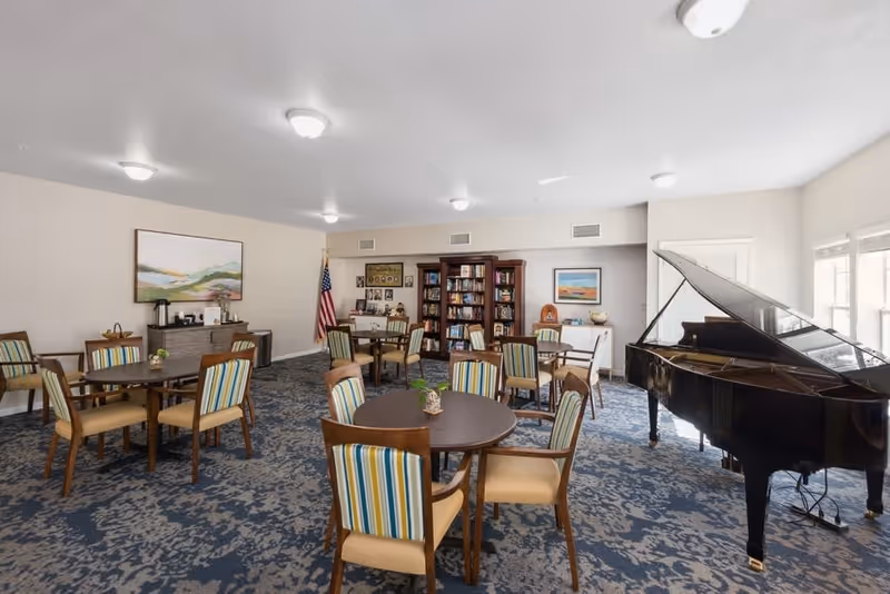 A spacious senior living common area with multiple round tables and chairs featuring striped upholstery. There is a grand piano on the right side near large windows allowing natural light. The back wall has a bookshelf filled with books, an American flag, and framed artwork. The room has a patterned carpet and ceiling lights.