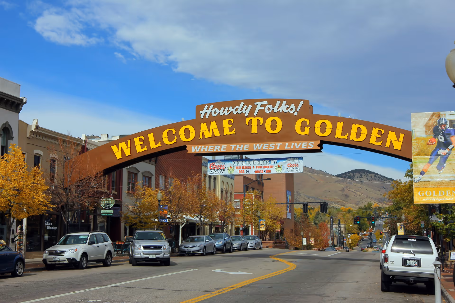 A street view in a small town with a large arching sign over the road that reads 'Howdy Folks! Welcome to Golden Where the West Lives'. Cars are parked along the street with buildings and trees showing autumn colors. Hills are visible in the background under a partly cloudy sky.